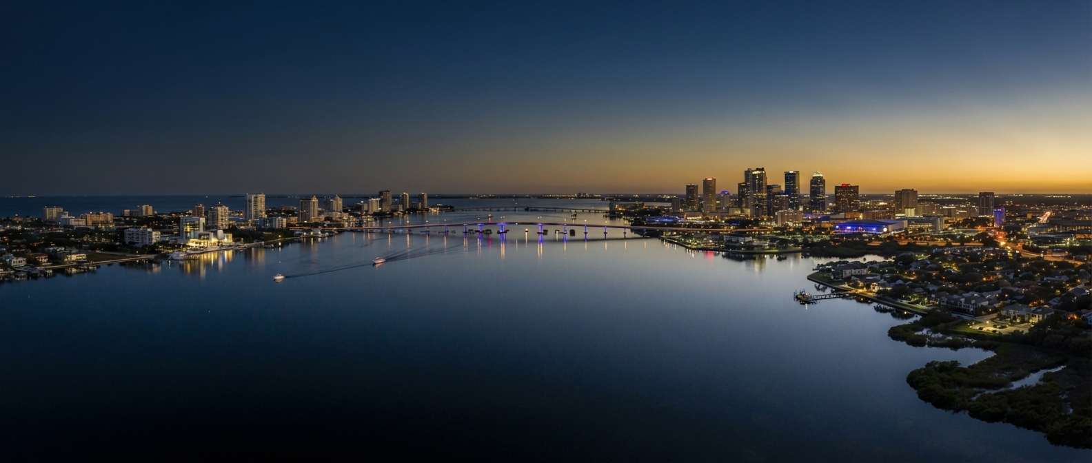 Editorial cinematic aerial of Tampa Bay at dusk — downtown Tampa and St. Petersburg mirrored in the bay in navy and gold tones